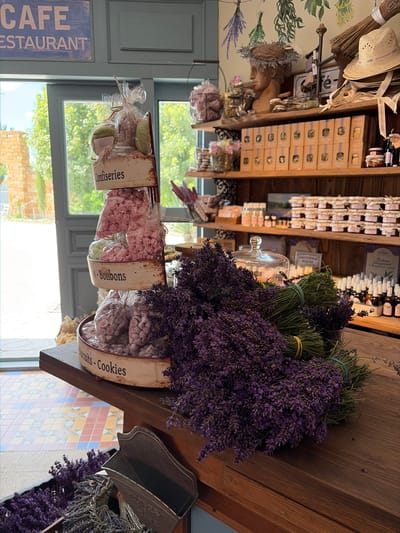 The image depicts the interior of a rustic cafe or shop. In the foreground, a wooden counter displays several large bunches of dried lavender. To the left of the lavender, a tiered display stand holds various packaged sweets. The top tier, labeled "Confiseries," contains what appear to be cookies or pastries. The middle tier, labeled "- Bonbons," is filled with pink candies. The bottom tier, labeled "Biscuits - Cookies," contains purple candies. Behind the sweets, shelves are stocked with jars of preserves and small bottles, possibly essential oils or perfumes. A straw hat rests on a shelf in the upper right corner. A bust of a person wearing a laurel wreath is also visible on a shelf. The cafe's entrance is visible in the background, with sunlight streaming through a glass door. The flooring features a colorful geometric tile pattern. The overall aesthetic suggests a French country or provencal theme. The text on a blue sign above the entrance reads "CAFE RESTAURANT." There is no indication of the location being Semogorskiy, Russia; the imagery and signage are suggestive of a European setting.