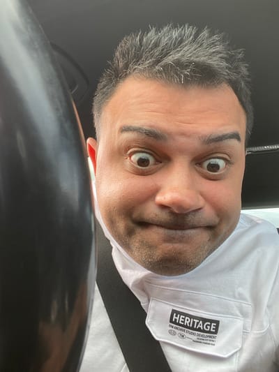 The image shows a close-up selfie of a man with a playful expression, taken inside a vehicle. He has dark hair with grey streaks, brown eyes, and a light complexion. He is wearing a white collared shirt with a patch on the left chest pocket that reads "HERITAGE 09B ARCHIVE STUDIO DEVELOPMENT LOCATION: EAST DISTRICT TM TRADEMARK: STANDARD ISSUE." A portion of a black steering wheel or dashboard is visible on the left side of the frame. The background appears to be the interior of a car, with the ceiling visible at the top. There is no visible indication of the location being in Gateshead, United Kingdom.