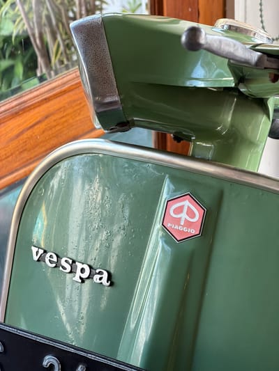 A close-up shot focuses on the rear section of a vintage green Vespa scooter. The scooter displays the "Vespa" logo in silver lettering on its side panel, along with a hexagonal Piaggio badge in red and white. The paintwork shows signs of age, with some bubbling and wear. A chrome trim runs along the edge of the panel. In the background, hints of greenery and wooden textures suggest an outdoor or indoor setting with natural elements. The scooter appears to be parked, with a black license plate partially visible at the bottom of the frame. The image likely depicts the scooter in its current state in Pekutatan, Indonesia, without any specific actions or interactions taking place.