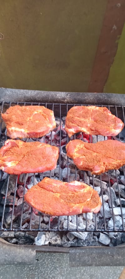 A close-up, overhead view shows five seasoned cuts of meat on a metal grill rack. The meat, which appears to be pork, is coated in a reddish-brown spice rub. The grill is positioned over glowing charcoal embers, with some heat visible as red and orange hues through the gaps in the grate. The grill itself is dark and appears to be made of metal with a textured handle visible on the right side.
To the left of the grill, the background is a mottled greenish-brown surface, possibly a wall or tarp, with a rougher, reddish-brown band running along the top edge. To the right of the grill, a speckled gray surface is visible, reminiscent of concrete or a patterned tiled floor.
The scene suggests an outdoor cooking event, likely a barbecue. The lighting indicates it could be daytime. No people are visible in the frame. The overall impression is one of preparing food for grilling. The context provided places the scene in Iskrets, Bulgaria.