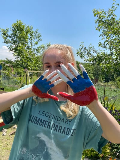 In the city of Yasnoe, Russia, a young woman with blonde hair stands in a garden on a sunny day. She is wearing a teal t-shirt with the text "LEGENDARY SUMMER PARADISE RELAX & SURF" printed on it. Her hands are painted with the colors of the Russian flag: red on the palm and lower fingers, blue in the middle, and white on the fingertips. She is holding her hands up in front of her face, partially obscuring it, as she looks at the camera. The background shows lush green trees, a fence, and a clear blue sky, suggesting an outdoor recreational area.