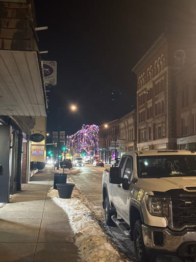 The image captures a winter night scene on a city street in Leominster, United States. The perspective is from a slightly elevated position looking down a street lined with buildings.
In the foreground to the right, a large white pickup truck, possibly a GMC, is parked on the side of the street. Its chrome grille and headlights are prominent. A significant amount of snow is piled along the curb and on the sidewalk next to the truck, indicating cold weather conditions. The sidewalk in front of the truck is cleared.
Across the street, a large tree is heavily adorned with numerous colorful Christmas lights, creating a vibrant focal point. Streetlights illuminate the scene, casting a warm glow on the ground and buildings. Several traffic lights are visible further down the street, one showing a green signal.
On the left side of the street, a row of older brick buildings with intricate architectural details lines the block. These appear to be multi-story commercial or residential structures, typical of a downtown area. Faint, partial text "Henson" is barely discernible on a sign on one of these buildings. Further down, a circular sign with a logo and the word "Store" is visible, along with other blurry commercial signs, including one that might read "Colonial" and another with blue and red text.
The overall atmosphere is festive and quiet, characteristic of a winter night during the holiday season. The absence of people on the street contributes to a sense of stillness.