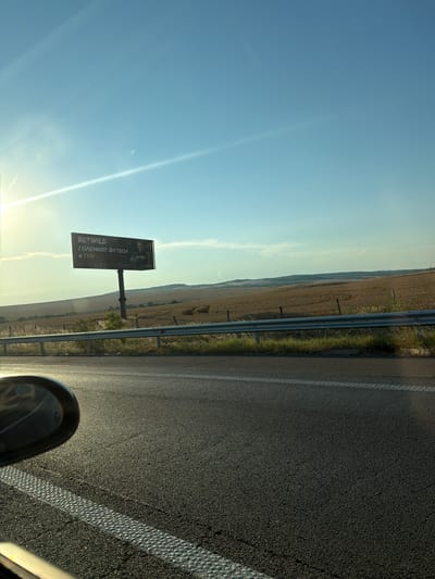 The media file depicts a road scene in Troyanovo, Bulgaria, likely during the late afternoon or early evening, given the low angle of the sun and the warm, golden light. The camera is positioned at a slightly oblique angle, looking forward and to the side, suggesting it was taken from a moving vehicle.
The main subjects include a stretch of asphalt road with a white line marking its edge, and a guardrail separating the road from a rural landscape. Beyond the guardrail, fields of dry, golden crops stretch towards rolling hills in the distance. The sky is a clear, pale blue with a few wispy clouds.
A prominent object in the scene is a billboard mounted on a pole. The billboard displays an advertisement, and visible text on it reads: "BETWILD," "ГОЛЕМИЯТ ФУТБОЛ," and "e ТУК." The "BETWILD" text is in English, while the other phrases are in Cyrillic, suggesting Bulgarian language. The advertisement appears to be for a betting company, likely related to football.
A notable detail is the lens flare caused by the sun, which creates bright streaks of light across the sky and the scene. The overall mood is one of travel and openness, with the vast rural landscape and the clear sky evoking a sense of journey. The presence of the road and the billboard place the scene firmly in a roadside setting. No people are directly visible.