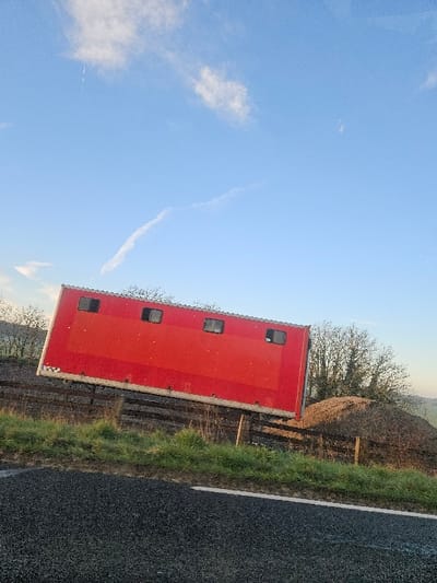 The image depicts an outdoor scene in Émancé, France, under a clear blue sky with scattered white clouds and a faint contrail. A large, red, rectangular trailer, consistent with a horse or livestock trailer, is positioned diagonally in the midground, with its left end lower than its right. The trailer's visible side features four small, square, dark-framed windows or vents. It appears to be resting on or immediately behind a wooden fence constructed of horizontal rails and vertical posts. In the immediate foreground, a dark asphalt road with a continuous white line marking its edge extends horizontally across the bottom of the frame. A strip of green grass and dry vegetation is situated between the road and the fence. Behind the right side of the trailer and the fence, a substantial mound of dark brown, granular material, likely soil or compost, is visible. Deciduous trees with sparse foliage or bare branches are present in the background, particularly on the left and right, against a distant landscape of gentle hills. No individuals are present in the scene, and no active actions are depicted.