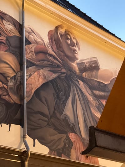 The image shows a mural painted on the side of a building in Deventer, Netherlands. The mural depicts a person from the chest up, wearing a headscarf and draped in richly patterned fabrics. The person's face is turned upwards and to the right, with their eyes closed or looking down, conveying a sense of contemplation or perhaps weariness. They appear to be carrying a rectangular object wrapped in fabric on their shoulder. The style of the mural is realistic, with soft lighting and detailed rendering of the textures and folds of the clothing. The mural is partially obscured by a modern awning or overhang in the foreground, which casts a shadow on the lower right portion of the artwork. The background of the mural is a light, neutral color, suggesting a painted sky or backdrop. The building itself appears to have traditional architectural elements, with a dark roofline visible at the top of the image. A downspout runs vertically along the left side of the mural.