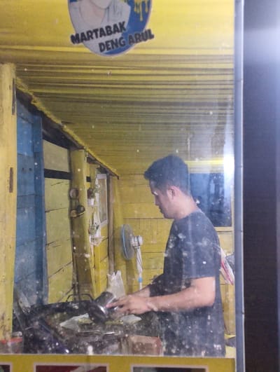 The image depicts a man in a black t-shirt working at a food stall in Pompanua, Indonesia. The stall has a yellow corrugated metal roof and walls, with some blue wooden panels visible. A sign above the stall reads "MARTABAK DENG ARUL". The man is standing behind a counter that appears to be made of dark metal, with various cooking utensils and ingredients visible. A white fan is mounted on the wall behind him. The scene is reflective, suggesting the image was taken through a glass surface, possibly a window or a display case, which has smudges and speckles on it. The overall environment appears to be an outdoor, perhaps street-side, food vendor setting.
