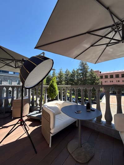 The image depicts an outdoor seating area on a balcony or patio, likely in the city of Jermuk, Armenia, as suggested by the architecture and landscape. In the foreground, a large, octagonal softbox on a stand is positioned to the left, suggesting a photography setup. To the right, there is a dark grey, oval-shaped table with two small items placed on its surface: a black camera lens and a slim, rectangular object that appears to be a cigarette lighter or a card case.
Behind the table, a white sofa with woven rattan sides and a light-colored cushion is visible. The balcony itself is paved with dark wooden planks. A decorative stone balustrade with ornate columns runs along the edge of the balcony, separating it from the area below. Beyond the balustrade, a row of tall, green coniferous trees forms a backdrop. In the distance, a multi-story building with a light pink facade and dark windows is visible, along with parked cars. The sky is clear and blue, indicating a sunny day. Two large, beige cantilever umbrellas provide shade over the seating area. The overall scene suggests a quiet, well-appointed outdoor space, possibly for relaxation or a photoshoot.