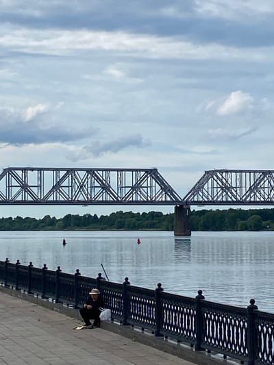 The image, displayed horizontally, captures a tranquil river scene under a cloudy sky. A substantial steel truss bridge dominates the midground, spanning across a wide body of water. The bridge exhibits intricate geometric patterns in its structure, supported by a sturdy pier positioned in the river.

On the far side of the river, a dense line of green trees marks the riverbank. The river's surface appears calm, reflecting the muted light of the sky, with two small red buoys visible. In the foreground, a stone embankment with a dark, ornate railing stretches along the near side of the river.

A person, wearing a hat and dark clothing, is seated on the embankment near the railing, appearing to be fishing or observing the water. A fishing rod extends from their position towards the river, and a white bag or other personal items are placed beside them. The time of day is daylight, characterized by an overcast or partly cloudy sky. The setting suggests an outdoor, perhaps leisurely, activity in Grigor'evskoe, Russia. No visible text is present in the image.