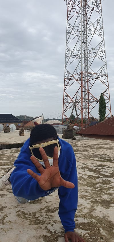 The media file displays a person in the foreground, wearing a blue hoodie and white sneakers. They are positioned on a textured, light-colored surface, possibly a rooftop, with a large, metallic tower structure dominating the background. The tower has a complex lattice design, with sections painted white and reddish-brown. In the distance, several buildings and trees are visible. The sky is overcast with clouds, suggesting a daytime setting with potentially dim lighting. The person is holding their hand out towards the camera, palm facing forward, in a gesture that could be interpreted as a greeting or a request to stop. Their face is partially obscured by large, white-framed glasses. The overall scene appears to be outdoors, in a somewhat industrial or urban environment, given the presence of the tower and buildings. The location is identified as Anguldi, Nigeria. There is no visible text in the image.