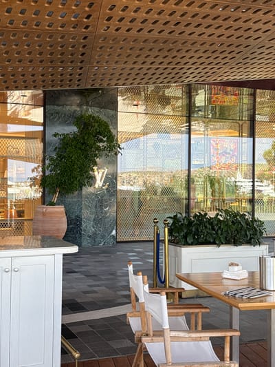 The image depicts an outdoor seating area in front of a Louis Vuitton store. The store features a facade of green marble with a prominent gold Louis Vuitton logo. A large potted ficus tree stands to the left of the entrance. The outdoor area has a wooden deck with a white cabinet on the left and a wooden table with white canvas chairs on the right. The table is set with a menu and what appears to be sugar packets. In the background, a reflective glass building with intricate metalwork is visible, reflecting the surrounding landscape and artwork. The scene is set during the daytime, with clear sunlight casting shadows. The location is identified as Vouliagmeni Municipal Unit, Greece.