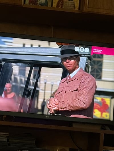 The image shows a television screen displaying a scene from a program. A man wearing a black hat, glasses, and a pink and white striped jacket is prominently featured. He is looking towards the viewer. To the top right of the screen, there is a graphic with the letters "тет" and Ukrainian text that translates to "in love with life." In the background, partially visible through the window of a vehicle, is a person with short, light-colored hair. The setting appears to be urban, with buildings and a yellow and red graphic of a rooster visible in the background. The specific location in the city of Osolynka, Ukraine, cannot be determined from the visual information provided.
