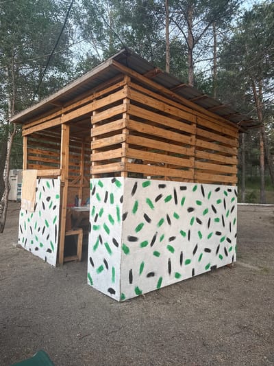 This media file captures an outdoor scene in Zerendi, Kazakhstan, featuring a rustic, open-sided wooden structure. The structure, likely a shed or booth, is built with a frame of rough-cut timber or repurposed pallets and is topped with a corrugated metal gabled roof.
The front and one visible side of the structure are clad with white panels, which are decorated with a repeating pattern of short, brushstroke-like dashes in alternating green and black. The left side of the structure remains open, revealing its wooden slat construction. Inside, some indeterminate objects are visible, including what appears to be a wooden box or table. The ground surrounding the structure is composed of gravel and dirt. A partial view of a green plastic chair is seen in the foreground to the right.
The setting is a wooded area, with numerous tall conifer trees, likely pines, forming a dense canopy in the background and overhead. Patches of blue sky are visible through the foliage. The natural light indicates it is daytime, with soft illumination suggesting late morning or early afternoon.
No people are present in the image, and no specific activity is taking place. A small, light-colored piece of material, possibly paper, is affixed to the lower front painted panel. Faint Cyrillic text can be discerned on this material, including fragments like "МИР" (meaning "world" or "peace") and "СТРОИТЕЛЬСТВО" (meaning "construction"). The structure has a handcrafted appearance, blending with its natural surroundings.