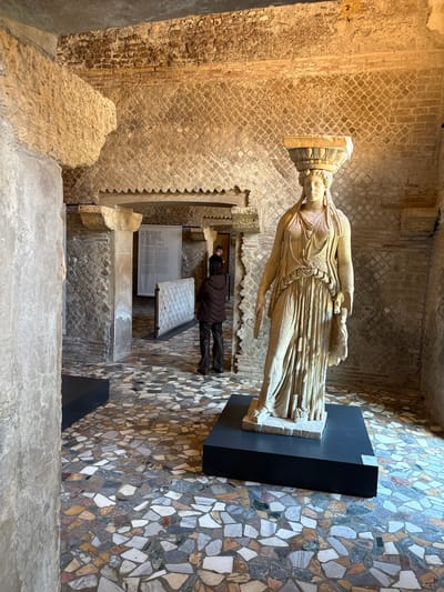 The image shows an interior space in the ruins of Hadrian's Villa in Tivoli, Italy. A classical statue of a woman on a black display stand is centrally located. A person wearing a dark jacket and pants is seen in the background looking at a display, partially obscured by architectural elements. The floor is a mosaic pattern of small, irregular stones. The walls consist of different textures and materials, including brick and stone work. The overall lighting is soft and diffused.