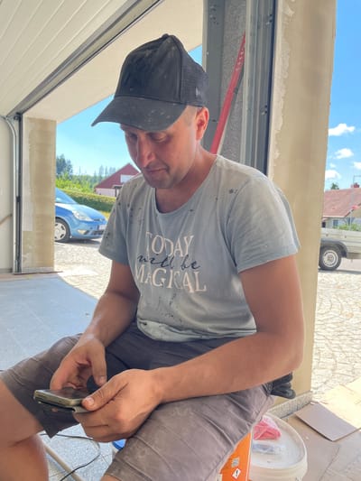 The image shows a man wearing a grey t-shirt with "TODAY will be MAGICAL" printed on it in a handwritten font, and a black baseball cap. He appears to be in the process of working on an outdoor construction or renovation project, possibly near a garage door. He is holding a mobile phone in his hands and looking down at it. His t-shirt and shorts are splattered with what appears to be paint or plaster, indicating he is actively involved in manual labor.

The setting is outdoors, under a bright, sunny sky with scattered clouds, suggesting it is daytime, likely late morning or afternoon. In the background, there is a blue car parked on a paved area, and glimpses of houses with red roofs and greenery, characteristic of a residential area. Construction materials, such as cardboard boxes and a white bucket, are visible nearby. The overall impression is a candid shot of a man taking a brief break from his work to check his phone. The location is identified as Jablonné nad Orlicí, Czechia.