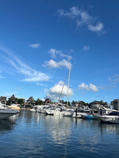 An outdoor, eye-level, vertical-format, medium shot captures a serene marina on a clear, sunny day. The sky is a vibrant blue, dotted with wispy white clouds. Below, a calm body of water reflects the sky and the numerous yachts docked along a pier. The yachts vary in size and design, with white hulls and sleek profiles. In the background, a row of houses with distinctive, multi-tiered roofs, reminiscent of Thai architecture, are nestled amongst lush green trees. The overall scene evokes a sense of luxury and tranquility in a tropical setting. The location is identified as a marina in Ko Kaeo, Thailand.