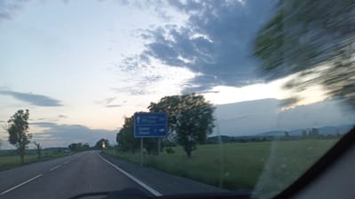The image depicts a view from inside a vehicle traveling on a road in what appears to be the outskirts of Sokolovo, Bulgaria. The vehicle is moving, as evidenced by motion blur. A road sign indicates "София A26" (Sofia A26) pointing upwards and "Кюлевча 5" (Kolevcha 5) pointing right. The surrounding environment includes green fields, trees, and distant mountains. The sky is cloudy, suggesting either dawn or dusk. No people are visible inside or outside the vehicle.