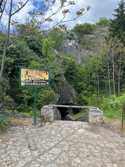 The image shows the entrance to Magura Cave, near Raianovtsi, Bulgaria. The entrance is framed by stone blocks and a metal grate. A sign for "Magura Cave" is positioned nearby. The area is vegetated with trees and bushes. The background features a rocky cliff. The foreground is a stone path. The sky is partially cloudy.