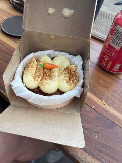 The image shows a small round cake topped with white frosting and sprinkled with cinnamon in a cardboard box. The cake is sitting on a wooden table. A red aluminum can with white writing is visible in the background. The scene is taken from a perspective above the cake. The cake appears to be freshly baked, and the cinnamon sprinkles are evenly distributed across the surface. The can is partially obscured by the edge of the table. The location is the city of Majadahonda, Spain. The coordinates are: 40.469, -3.857.