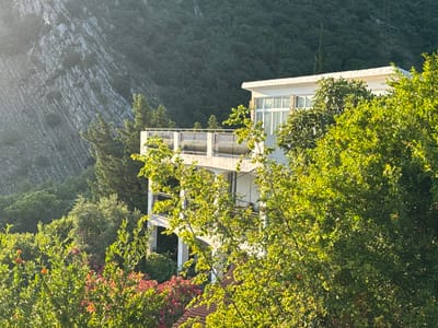 The image depicts a multi-story white building nestled amidst lush green foliage and a rocky mountainside in Čučuci, Montenegro. The building features balconies with decorative railings and large windows. Several trees, some with bright green leaves and others with pink flowers, surround the structure. The rocky mountain, covered with sparse vegetation, forms the backdrop. The scene is bathed in the warm, soft light of what appears to be late afternoon or early morning. No people are visible in the image.