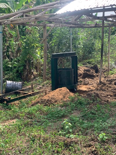 A dark green compost bin with a metal frame and a metal drum is situated in a small, unpaved area in a backyard in Egbeda, Nigeria (latitude: 7.12, longitude: 5.362). There is a pile of dirt next to the bin. Behind the bin is a tall, leafy, green tree. There is a wooden structure with a corrugated metal roof. There are green plants growing in the area. The ground is covered in dirt and leaves. The bin is tilted slightly to the right. The metal drum is partially open, and a small amount of dirt is visible inside. The area appears to be relatively unused, with signs of minimal activity.