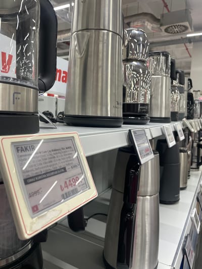 This media file captures a retail display of kitchen appliances, specifically tea makers, in what appears to be a store in Bağcılar, Turkey. The scene is a close-up, angled shot showcasing rows of sleek, stainless steel tea makers with black accents. Several of the appliances have digital price tags attached, displaying prices in Turkish Lira. One prominent tag shows a price of "€4490" and another indicates "549,50" as a protection insurance cost. Text on the tags includes brand names like "FAKIR" and "TEFAL," along with product specifications such as "TEA_MAKER," "3.2 kg," and "21 cm." The overall impression is one of commercial display, highlighting the products and their pricing. The lighting suggests an indoor setting, possibly a department store or electronics retailer. There are no people visible in the frame.