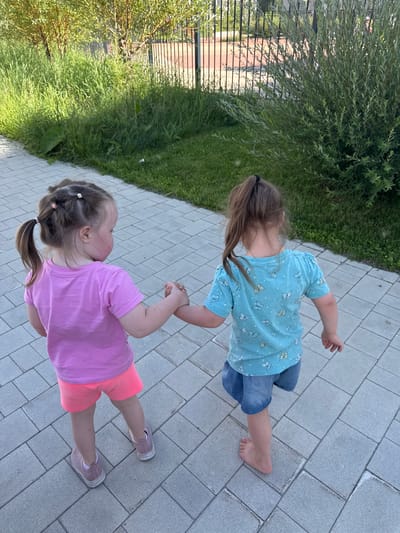 Two young girls are walking on a paved walkway. The girl on the left, facing away from the camera, wears a pink t-shirt and orange shorts, with her hair in two pigtails. The girl on the right, also facing away, wears a blue t-shirt with a butterfly pattern and denim shorts, with her hair in a single ponytail. They are holding hands. The walkway is made of light gray rectangular pavers. To the left of the walkway is a grassy area with tall green grass and trees, and to the right is a dense green shrub. In the background, a black metal fence encloses a reddish-brown surface, possibly a sports court. The scene appears to be in an outdoor, urban or suburban environment.