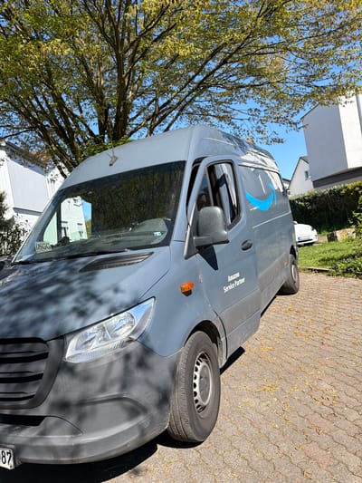 An Amazon Service Partner delivery van is parked on a paved driveway in Maintal, Germany. The van is gray with the Amazon logo on the side. A tree with budding leaves is visible behind the van. The location is residential, with houses visible in the background. The vehicle license plate indicates registration in Germany. The cobblestone driveway is littered with dried blossoms.