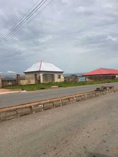 The image shows a street scene in Kona Garu, Nigeria. The perspective is from a slightly elevated angle, looking down towards the road and adjacent buildings. The sky is overcast with fluffy white clouds, suggesting a daytime scene.
On the left side of the image, there are power lines running diagonally across the frame. Further to the left, partially obscured by the power lines, are more buildings, one with a distinctive red roof. In the foreground, there is a building with a white and red corrugated metal roof, constructed with stone walls. It appears to be a residential or commercial building, with barred windows and a metal gate. Behind this building, a sign is visible, attached to a fence. The text on the sign reads "FOUNDATION ACADEMY INT'L." The surrounding area is covered with grass.
The road occupies the right side of the image and is paved with asphalt. It is bordered by a raised kerb made of concrete blocks. The road surface shows some signs of wear and tear, with darker patches and a few small white objects that might be litter.
The overall impression is a typical street view in a Nigerian town, with a mix of residential and possibly educational buildings along a main road. The cloudy weather adds a muted quality to the scene. There are no people visible in the image.