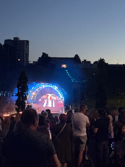 The image shows an outdoor evening event in Jonava, Lithuania, likely a concert or festival, with a stage featuring a large arched screen illuminated with blue and white lights. A man is visible on the screen, appearing to be a performer or speaker. A crowd of people is gathered in front of the stage, facing the screen. Some trees are present in the foreground and background. In the far background, buildings are visible against the twilight sky. The scene suggests a lively public gathering.