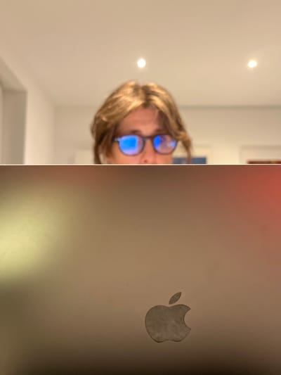 An indoor scene is depicted from a low, upward-angled perspective. In the upper portion of the frame, the blurred upper head of an individual with medium-brown hair is visible, looking forward. The individual wears dark-framed eyeglasses, the lenses of which display prominent blue reflections, indicating anti-reflective or blue light filtering properties. Below the individual's head, occupying the lower half of the image, is the metallic silver or gray back casing of an open laptop. A distinct, light gray Apple Inc. logo is centrally located on the lower right section of this casing. The laptop surface exhibits minor variations in tone and texture. The background, largely out of focus, shows a white ceiling featuring two recessed, warm-toned spotlights. No visual cues within the image enable the determination of its specific location within the city of Foetz, Luxembourg, or any other geographical area.
