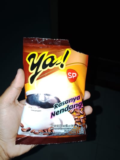 A hand is holding a small, torn packet of coffee. The packet is predominantly yellow and orange, with a large black and yellow "Ya!" logo at the top. Below the logo, the text "Rasanya Nendang" is printed in pink and purple. There's an image of a coffee cup with milk and coffee beans. Text on the packet indicates "Berat Bersih : 60 g", "Kopi Campur", and "HALAL". The packet is slightly damaged at the top. The background is dark and out of focus, suggesting an indoor or dimly lit environment. No specific location in Sendangrejo, Indonesia, is depicted.