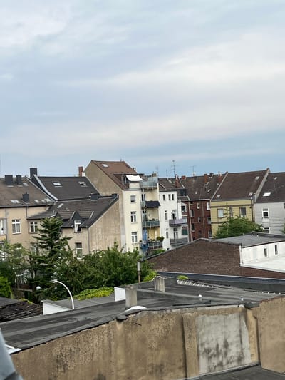 This image captures a view of urban rooftops and buildings in Neuss, Germany, under an overcast sky. In the foreground, the rough, textured surface of a beige wall with greyish, mottled discoloration dominates the bottom portion of the frame. Above this wall are several flat, dark grey rooftops, one of which has visible white vents or caps. To the left, a curved white lamp post is visible above another section of flat rooftop.
Behind the foreground rooftops, a dense cluster of green trees and foliage partially obscures the lower levels of buildings. These buildings are diverse in their architectural style and color. Several multi-story residential buildings with tiled roofs of varying shades of brown and grey are visible. Some have visible chimneys and dormer windows. The building facades are painted in muted tones of beige, yellow, and brick red. Windows and balconies are present on these structures. In the far background, the sky is a pale, uniform grey, indicating cloudy weather. No people or distinct actions are captured in this scene.