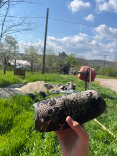 A person's hand holds a JBL Charge 5 speaker with a camouflage pattern. The speaker is the prominent object, and the background appears to be a yard in the city of Smilets, Bulgaria. There is green grass, trees, and a building in the background. A red cement mixer is also visible. The sky is blue with clouds.