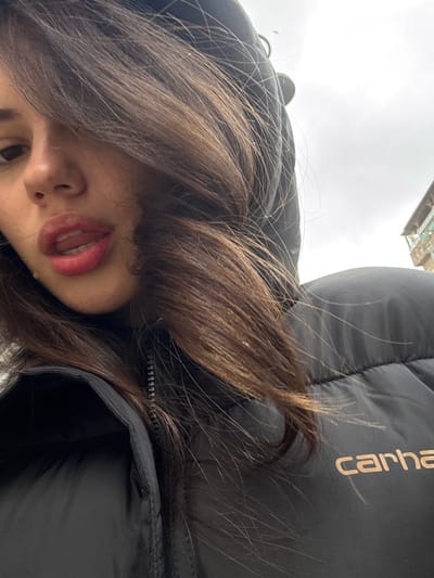 A close-up, slightly upward-angled image captures a female individual with dark brown hair styled to the side, partially obscuring her face. Her left eye is visible, and her lips, coated with red lipstick, are slightly parted, revealing the tip of her tongue and upper teeth. She wears a dark, likely black, puffer jacket featuring a visible zipper and an indiscernible hood pulled over her head. A light brown "carhartt" logo is discernible on the wearer's right side of the jacket. The background consists of an overcast, cloudy sky, with a section of an aged, multi-story building featuring a balcony visible in the upper right. The scene is located in Rehovot, Israel.