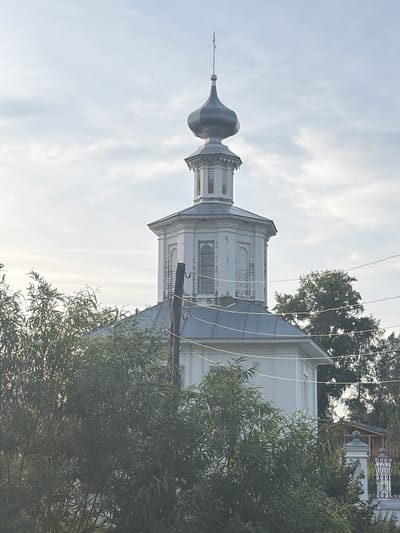The image shows the bell tower of the Church of the Resurrection in Cherdyn, Russia. The church is white with a gray roof and a dark blue dome topped with a cross. The bell tower is octagonal and has white walls with some visible brickwork. There are windows on each side of the tower. Trees and foliage obscure the lower portion of the church and bell tower. Power lines run horizontally across the image in front of the bell tower. The sky is overcast with some light clouds.