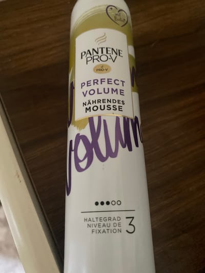 A white aerosol can for Pantene Pro-V Perfect Volume Mousse stands on a wooden surface. The can has a gradient of gold and white at the top, with purple lettering that spells out "VOLUME" in a large script across the middle. Below the large script, it reads "HALTEGRAD NIVEAU DE FIXATION 3" with three black dots preceding it. The can appears to be in focus, with the background of the wooden surface slightly blurred. The image is taken from a slightly elevated angle, looking down at the can.