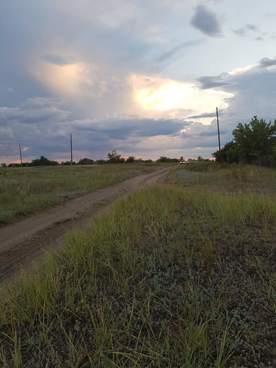 This image depicts a rural landscape in the Dolen Rural District of Kazakhstan. The scene is characterized by a dirt road cutting through a grassy field. The sky is filled with dramatic clouds, with the sun peeking through, suggesting either sunrise or sunset. A few scattered trees and utility poles are visible along the horizon, hinting at the presence of a nearby settlement. In the distance, a small vehicle can be seen driving on the road, adding a sense of movement to the otherwise tranquil setting. The overall mood is one of peacefulness and the beauty of nature, bathed in the soft light of the declining or rising sun. There is no visible text in the image.