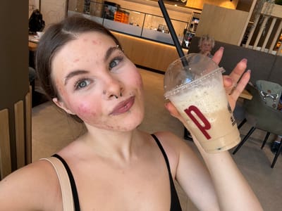 The image shows a young woman with acne and multiple facial piercings, holding a partially consumed iced coffee in a clear plastic cup with a black straw. The cup has a red "p" logo. She is wearing a black top with a beige strap visible on one shoulder. The background shows the interior of what appears to be a cafe, with light wood paneling, tables, and chairs. A reflection of an elderly woman is visible in the plastic cup. Based on the "p" logo and the general appearance of the cafe, the location is likely a branch of the "Peter Pane" restaurant chain, which has a location in Fulda, Germany.