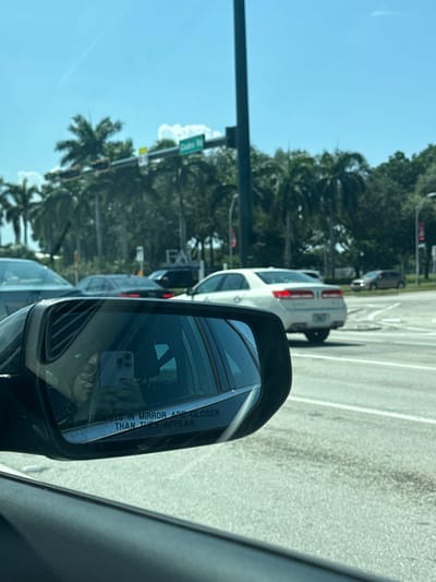 The image depicts a daytime street scene from the interior of a vehicle, looking through the front passenger window and into the driver's side mirror. The location is an intersection on Glades Road, near Florida Atlantic University, in Boca Raton, Florida, United States, evidenced by a partially visible green street sign overhead reading "Glades Rd" and the distinct letters "FAU" visible on a structure or sign in the background amidst dense trees.
The sky is clear and blue with scattered white clouds. In the immediate foreground, the dark frame of the vehicle's interior and the driver's side mirror are prominent. The mirror's reflective surface contains a partial view of a person's hands holding a light-colored smartphone, actively capturing the photograph, and the etched text "OBJECTS IN MIRROR ARE CLOSER THAN THEY APPEAR."
Several other vehicles are present on the road. A dark-colored sedan is positioned in an adjacent lane to the left. A white four-door sedan, with its red tail lights illuminated, is visible ahead and to the right, appearing to be turning left or waiting at the intersection's marked stop line. Further down the road to the right, a lighter-colored crossover or small SUV is also visible. The environment features extensive tropical vegetation, primarily palm trees, lining the roadways and background, typical of the region. A tall, dark utility pole supports traffic signals at the center of the intersection.
