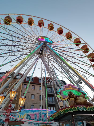 The image depicts a ferris wheel with illuminated gondolas and a sign reading "Original Berliner Riesenrad" at the top. The ferris wheel is adorned with colorful LED lights that create a vibrant spectacle against the clear blue sky. In the background, several apartment buildings are visible, suggesting an urban setting. At the base of the ferris wheel, a carousel-like structure is partially visible, featuring painted depictions of horses and an angel. The overall scene conveys a festive atmosphere, likely at a fairground or amusement park. The location is identified as Cottbus - Chóśebuz, Germany.