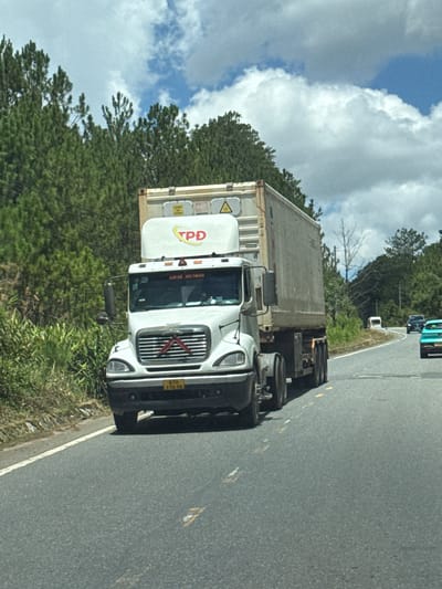 A white semi-truck with a grey shipping container is traveling on a paved road in Thôn 6, Vietnam. The truck is in the foreground and takes up a significant portion of the frame. It has a "TPD" logo on the side of the cab. The road appears to be a two-lane highway with a white dashed line marking the center and a solid white line on the right shoulder. Green trees and vegetation line the left side of the road. In the background, there are more trees on rolling hills, and a cloudy blue sky. Other vehicles, including a black car and a teal-colored car, are visible further down the road. The environment appears to be rural or semi-rural.