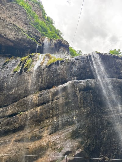 A low-angle, outdoor, full shot shows a rocky cliff face with a waterfall cascading down it. The cliff is dark gray and textured, with horizontal striations and patches of green moss growing in places. The waterfall appears as a shimmering veil of water, broken into several streams as it descends. Lush green trees are visible at the top of the cliff and on ledges. The sky above is overcast with thick, gray clouds. A thin, dark wire stretches diagonally across the upper portion of the frame. This image was taken in Khushto-Syrt, Russia.