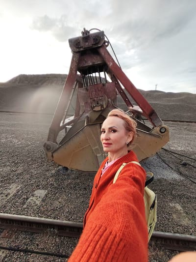 A medium shot captures a woman with short, pink-hued hair standing in front of a large, red and rusted industrial grabber or clamshell bucket. She is positioned on the right side of the frame, facing the camera and extending her right arm forward, suggesting she is taking a selfie. She wears an orange knitted cardigan over a pink top and a light-colored backpack with black straps. Her left ear is adorned with a heart-shaped earring. The background is dominated by the industrial machinery and piles of gravel or rocks. In the distance, a tall, slender structure, possibly a tower or mast, is visible on a hilltop under a cloudy sky. The ground in the foreground is covered in gravel and two parallel railway tracks run horizontally across the lower portion of the image. The overall environment suggests an industrial or quarrying location.
