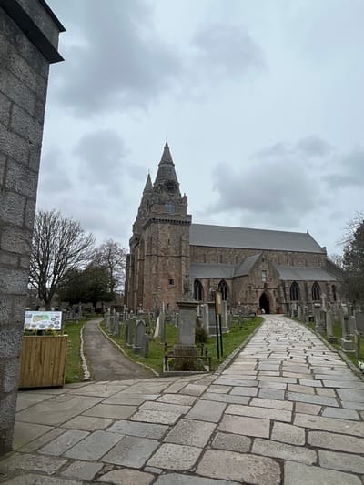 The image depicts St Machar's Cathedral in Aberdeen, Scotland. A stone pathway leads to the cathedral, lined with gravestones of the adjacent cemetery. The cathedral is made of grey stone, featuring a tall tower with a spire. The sky is overcast. A stone wall is in the lower left of the image. A sign stands near the pathway. Bare trees dot the scene.