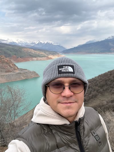 A male individual is prominently featured in a close-up photograph taken in Chorvoq, Uzbekistan. He wears a grey ribbed knit beanie with a "THE NORTH FACE" logo patch, dark-framed eyeglasses with amber-tinted lenses, an off-white hooded sweatshirt, and a dark olive puffer vest. The subject is looking directly at the camera with a neutral to slightly smiling expression. The background displays a wide body of light turquoise water, characteristic of the Charvak Reservoir, surrounded by hills and distant snow-capped mountains. The immediate left shows a reddish-brown, barren hillside with sparse, leafless vegetation descending towards the water. The sky is overcast.