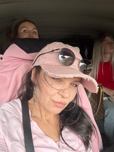Three women are seated in a vehicle. The woman in the foreground, wearing a pink baseball cap and sunglasses, is looking towards the camera. She has dark hair and is wearing a pink jacket. Behind her, to the left, another woman with brown hair is looking upwards, with only her eyes and forehead visible above a black collar. To the right, a woman with blonde hair wearing a red shirt is looking forward. A Louis Vuitton patterned bag is visible between the foreground woman and the woman on the right. The scene appears to be captured during daylight, with reflections of the outdoors visible in the sunglasses. There is no information in the image to suggest a specific location in Каменномостское сельское поселение, Russia.
