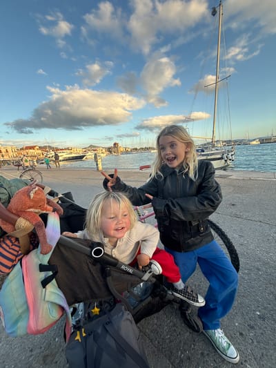 Two blonde children are featured in the foreground of a paved quay at the harbor of Aegina, Greece. An older child, approximately 8-10 years old, stands to the right of a dark-colored stroller, wearing a black leather jacket, blue pants, and light green canvas shoes. This child holds the right hand up in a 'peace' sign gesture and displays an open-mouthed smile revealing missing front teeth while looking towards the camera. Seated in the "ROYAL" brand stroller is a younger child, a toddler approximately 1-3 years old, wearing a light-colored knitted top, red pants, and plaid velcro shoes, also smiling towards the camera.

The stroller is laden with personal items, including a reddish-brown stuffed fox toy with a straw hat, a rainbow-colored backpack, and a dark gray backpack adorned with yellow star-shaped charms. A white bicycle is partially visible to the left, behind the stroller.

The background shows the harbor, filled with numerous moored sailboats and motor yachts on blue water. Along the distant waterfront, multi-story buildings with light-colored facades and terracotta roofs are visible, including a prominent white clock tower and a building with a distinct red domed roof. The sky above is blue with scattered white cumulus clouds, illuminated by warm, late-day sunlight. Distant hills are discernible on the horizon.