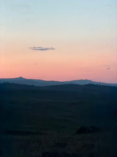 A scenic landscape photograph captures a serene twilight scene in Kuseevo, Russia. The sky transitions from a pale blue at the top to a soft pink and orange hue near the horizon, with a wispy cloud drifting across the upper portion. In the mid-ground, rolling hills are silhouetted against the colorful sky, with a distinct, pointed peak on the left and other fainter, rounded peaks further in the distance. The foreground is a dark, shadowy expanse of undulating terrain, possibly fields or moorland, with hints of vegetation visible in the lower part of the frame. The overall atmosphere is tranquil and picturesque, conveying the beauty of the natural landscape at dusk.