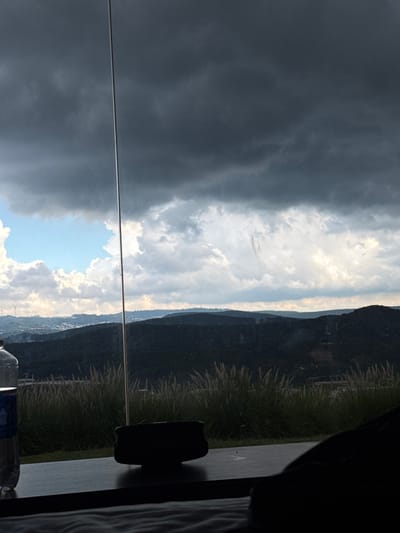 The image depicts a view from a window, looking out over a landscape under a dramatic, cloudy sky. In the foreground, a dark, rectangular object sits on a dark surface, possibly a table. To the left, a partially visible plastic water bottle is present. Beyond this immediate foreground, a dense field of tall, dry grass extends horizontally. The middle ground features dark, forested hills and a distant view of buildings and wind turbines, suggesting a semi-urban or rural setting. The sky is filled with heavy, dark clouds, with a small patch of bright blue sky and lighter, scattered clouds visible towards the center. The overall lighting is dim, indicating overcast conditions. There are no discernible people or specific landmarks that would indicate the location as Đa Lộc, Vietnam.