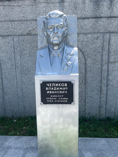A metallic bust of a man with stern features is positioned against a textured grey wall. The man is wearing a suit and tie, with several star-shaped medals pinned to his chest. Below the bust, a dark plaque displays text in Russian, identifying him as "CHELIKOV VLADIMIR IVANOVICH" and noting he is a "CAVALIER OF THE ORDER OF GLORY THREE DEGREES." The bust is mounted on a polished metallic pedestal, which extends downwards and is partially obscured by lush green grass and a grey stone surface. The overall scene is static and commemorative.